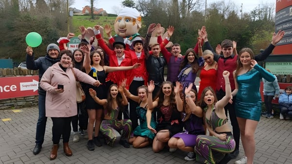 Un grupo de alumnas de la Kinsale Community School posan al aire libre frente a árboles y edificios en vestidos festivos coloridos.