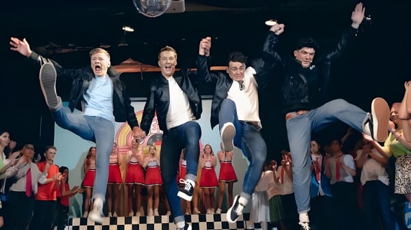 Un grupo de alumnas y alumnos de la Kinsale Community School está en un escenario con un patrón de tablero de ajedrez y un fondo decorado.