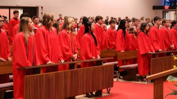 Estudiantes de la Kuemper Catholic High School llevan togas rojas y están sentados en un entorno similar a una iglesia.