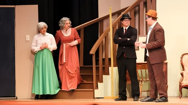 Estudiantes de la Kuemper Catholic High School presentan una obra de teatro en trajes históricos en un escenario con escalera de madera.