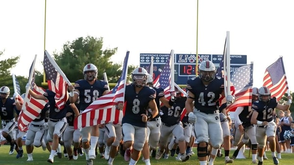 Un grupo de jugadores de fútbol americano corre con banderas estadounidenses hacia el campo de Lacey Township High School.