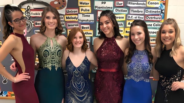 Un grupo de jóvenes mujeres en vestidos formales de colores posan frente a una pared con términos musicales en el terreno de Lacey Township High School.