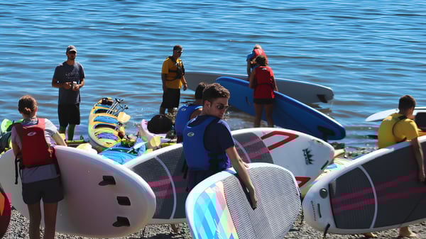 Estudiantes de la Ladysmith Secondary School están de pie en paddleboards en un cuerpo de agua con un fondo azul.