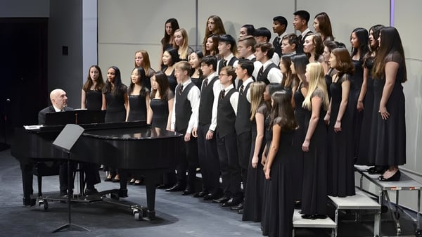 Un gran coro está de pie con ropa formal negra en el escenario de la Lake Forest Academy con un piano en primer plano.