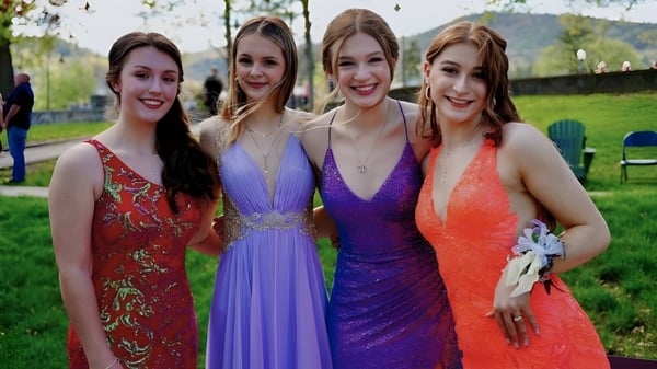 Cuatro alumnas de la Lake George Junior-Senior High School están juntas en vestidos coloridos en un prado con árboles de fondo.