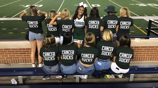 Un grupo de jóvenes mujeres con camisetas negras y el texto Cancer Sucks están en el campo deportivo de la Lake Orion High School.