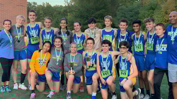 Un grupo de jóvenes atletas entrena en el campo deportivo de la Lake Ridge Academy con árboles y un edificio de ladrillo al fondo.