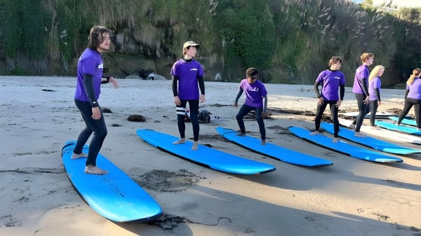 Estudiantes de la Lake Tahoe Preparatory School están de pie en camisetas moradas y trajes de neopreno en la playa junto a tablas de surf azules frente a un bosque verde.