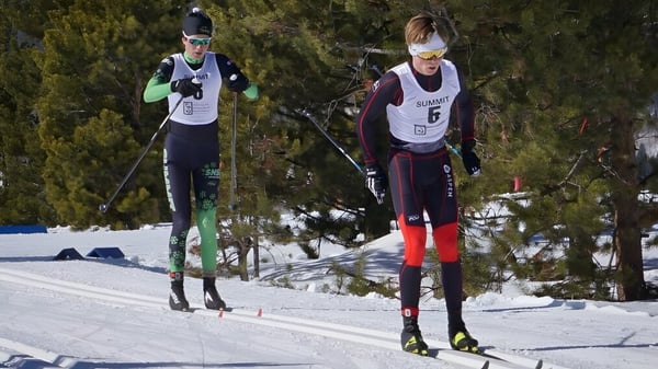 Dos estudiantes de la Lakes District Secondary School están esquiando en una pista nevada rodeados de pinos.