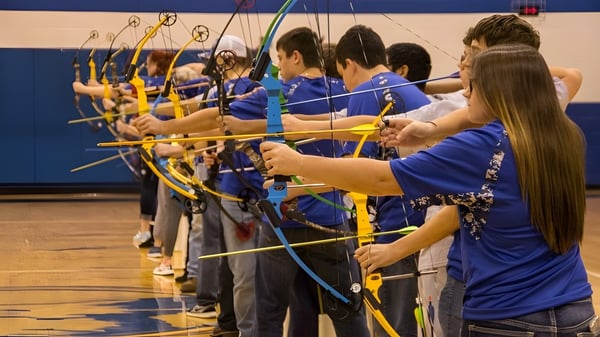 Estudiantes de la Lakes District Secondary School están en uniformes azules en el gimnasio y disparan con arco y flecha a dianas.