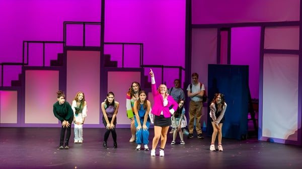 Un grupo de estudiantes de Lancaster Country Day School está de pie en un escenario frente a un colorido fondo morado y rosa con patrones geométricos.