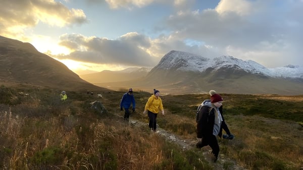 Un grupo de estudiantes del Lanesboro Community College camina por un sendero cubierto de hierba en un paisaje montañoso con picos nevados.