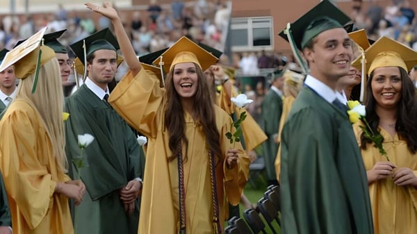 Graduados del Lanesboro Community College están al aire libre en togas verdes y doradas y saludan a la cámara.