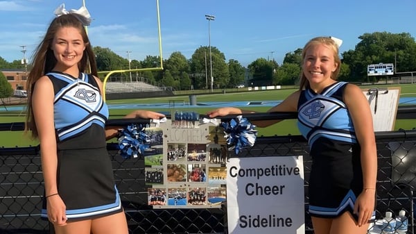 Dos animadoras en uniformes azules y blancos están en la banda de Lansing Catholic High School frente a un cartel deportivo.