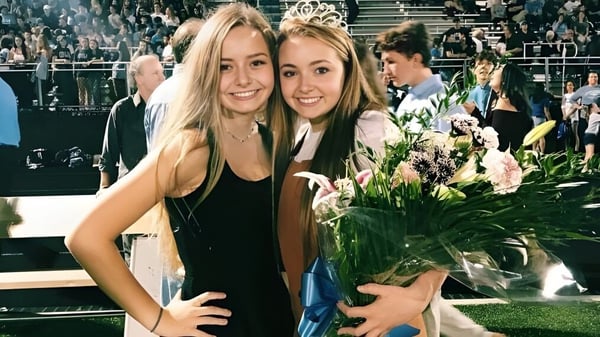 Dos estudiantes de Lansing Catholic High School sostienen juntas un ramo de flores y posan con una multitud en un escenario al fondo.