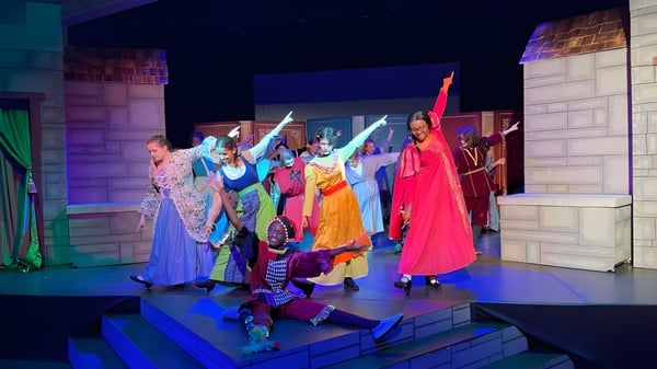 Un grupo de actores vestidos de colores se encuentra en el escenario de la Laurel School frente a un fondo arquitectónico con iluminación vibrante.