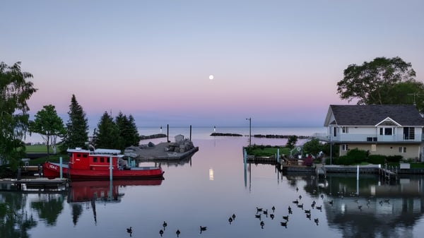 Un bote rojo está en la orilla del lago frente a casas y árboles en el terreno de la Leamington District Secondary School al atardecer.