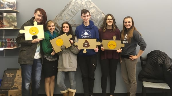 Un grupo de estudiantes de Leduc Composite High School posan con carteles coloridos en un aula con estanterías y obras de arte al fondo.