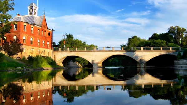 El edificio histórico de la École élémentaire et secondaire publique L'Équinoxe con torre del reloj está sobre un puente sobre un río tranquilo.