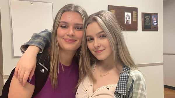 Dos estudiantes con cabello largo y rubio posan sonriendo en una sala con obras de arte enmarcadas en el campus de Lewisporte Collegiate.