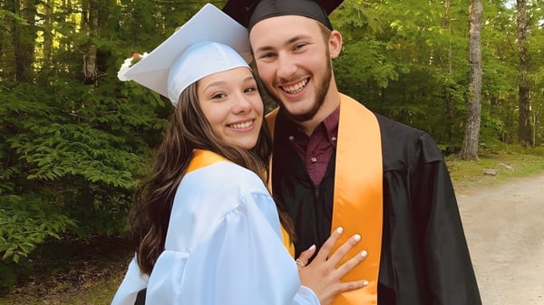 Dos graduadas de la Lincoln Academy en vestimenta de graduación están juntas en un área boscosa con abundante vegetación.