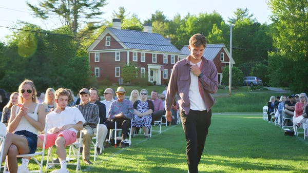 Un grupo de personas está sentado en sillas en una pradera frente a la casa de ladrillo rojo de la Lincoln Academy.