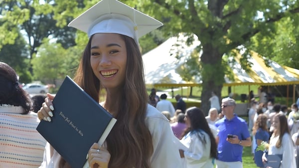 Una graduada de Linden Hall lleva birrete y toga y sostiene su diploma durante la ceremonia de graduación al aire libre.