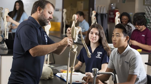 Un hombre está frente a un grupo de estudiantes de la Linfield Christian School sosteniendo un modelo de esqueleto durante la clase de anatomía.