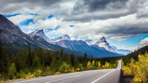 Una carretera sinuosa atraviesa un valle boscoso con montañas nevadas al fondo cerca de la Listowel District Secondary School.