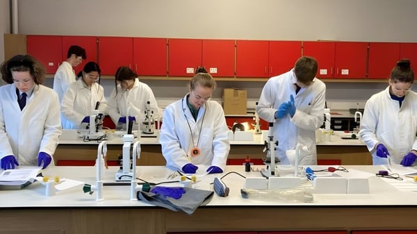 Estudiantes de la Longridge Towers School trabajan en batas de laboratorio blancas en diferentes estaciones en el laboratorio con equipo científico.
