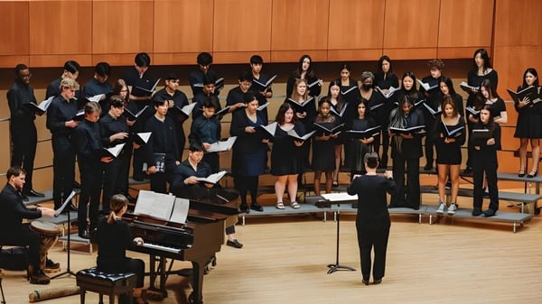 El coro de la Loomis Chaffee School está en túnicas negras en el escenario frente a una pared de paneles de madera con un piano en primer plano.