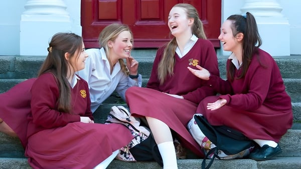 Las alumnas del Loreto College, St. Stephen's Green, están sonriendo con túnicas rojas frente a una puerta de madera roja.