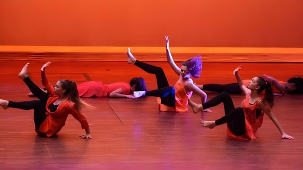Un grupo de estudiantes del Loreto College Cavan realiza un espectáculo de baile frente a un fondo de escenario naranja.