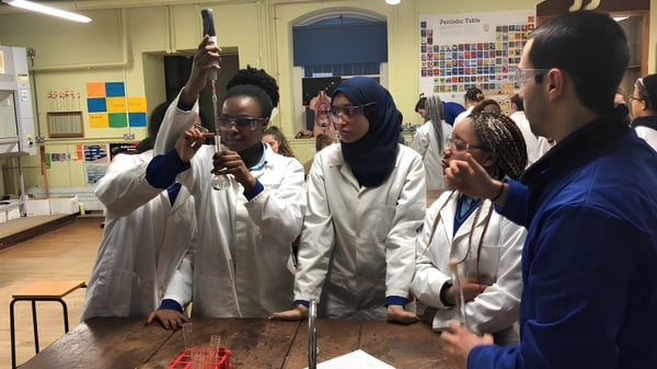 Estudiantes del Loreto College Mullingar trabajan en batas de laboratorio blancas en un aula con materiales científicos.