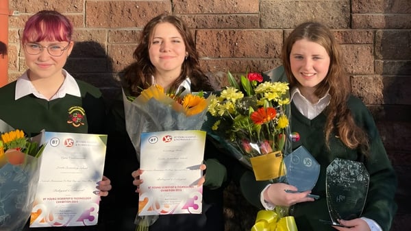 Tres alumnas de la Loreto Secondary School en Cork sostienen certificados y flores frente a una pared de ladrillo.