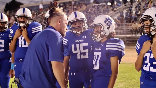 Un grupo de jugadores de fútbol americano en uniformes azules está en el campo del Los Angeles Unified School District con espectadores al fondo.