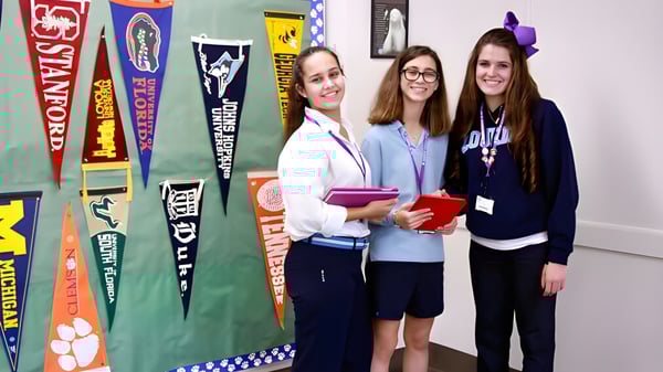 Tres estudiantes están en una sala con coloridos banderines escolares y pancartas en las paredes de Lourdes Academy.