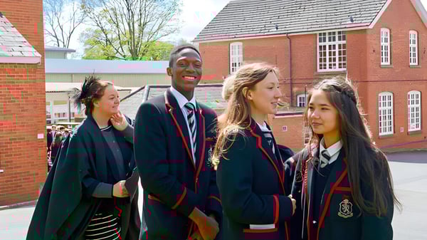 Un grupo de estudiantes en uniformes escolares está de pie y conversando frente al edificio de ladrillo de la Lucton School.