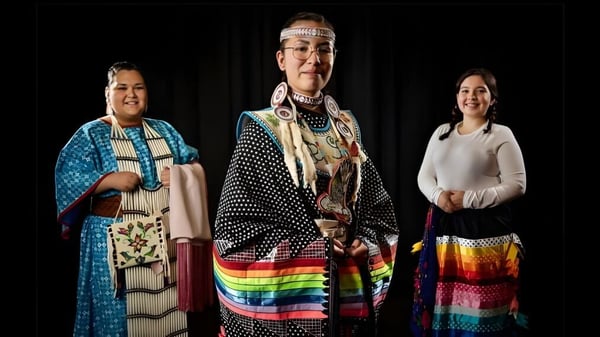 Tres personas con vestimenta tradicional indígena están de pie frente a un fondo oscuro en el campus de Luther College High School.