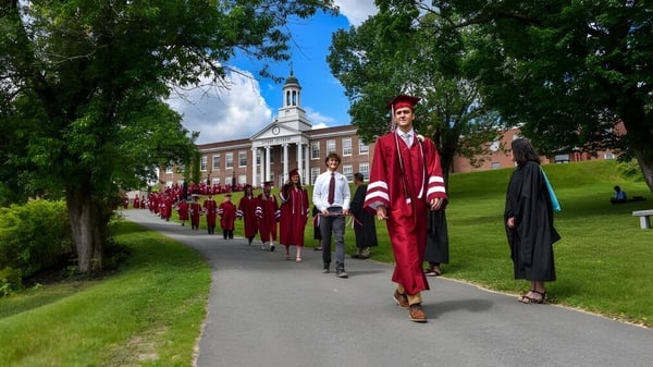 Graduados del Lyndon Institute con togas rojas caminan por un camino arbolado en el campus.