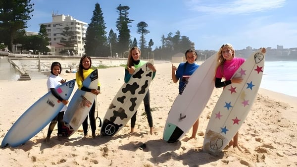 Alumnas del Mackellar Girls Campus están con tablas de surf coloridas en la playa frente a edificios y árboles.