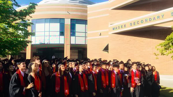 Un gran grupo de graduados y graduadas está frente al edificio de ladrillo de la Magee Secondary School con togas rojas y negras.
