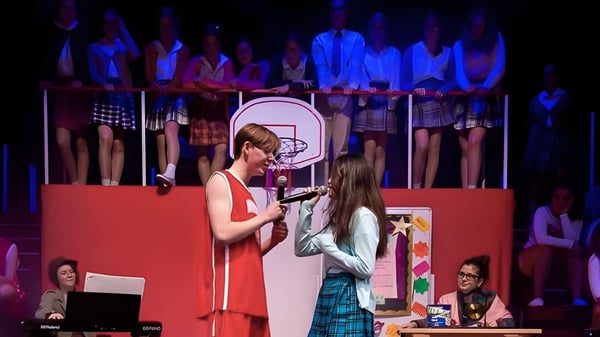 Una mujer con un vestido rojo y una niña con una falda a cuadros están en el escenario del Malvern College durante una actuación.