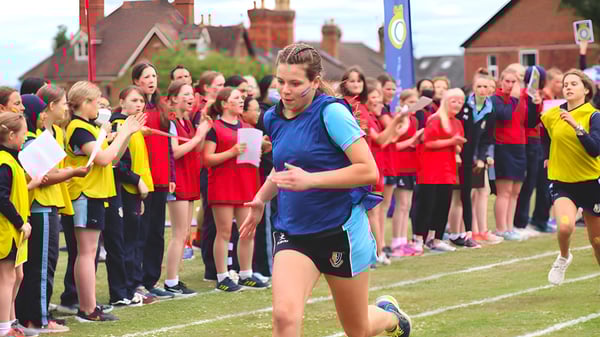 Un grupo de jóvenes atletas está en el campo de deportes de la Malvern St. James Girls' School con una mujer en un traje azul en primer plano.