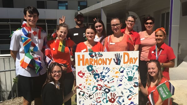 Un grupo de estudiantes del Manea Senior College sostiene una colorida pancarta con la inscripción Harmony Day frente a un edificio.