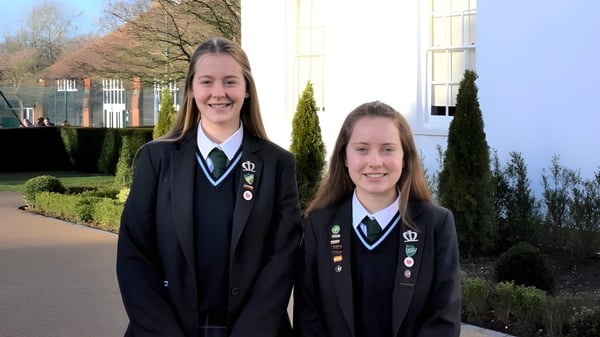 Dos estudiantes de la Manor House School están juntas afuera sonriendo frente a un complejo de edificios con árboles.