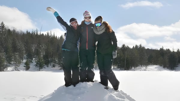 Tres personas con ropa de invierno caliente están de pie con los brazos en alto en un bosque nevado en el campus del Appleby College.