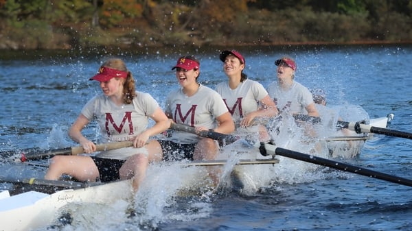 Cuatro estudiantes de la Marianapolis Preparatory School reman en un lago rodeado de vegetación otoñal.