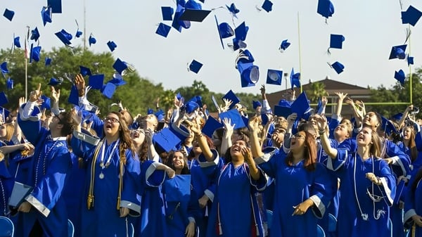 Un gran grupo de graduados de Marion County Public Schools lanza los birretes de graduación azules al aire frente a un fondo de árboles.