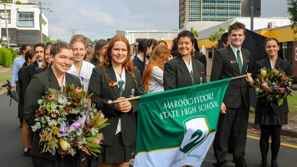 Un grupo de estudiantes de la Maroochydore State High School lleva togas de graduación y sostiene un banner frente a rascacielos.